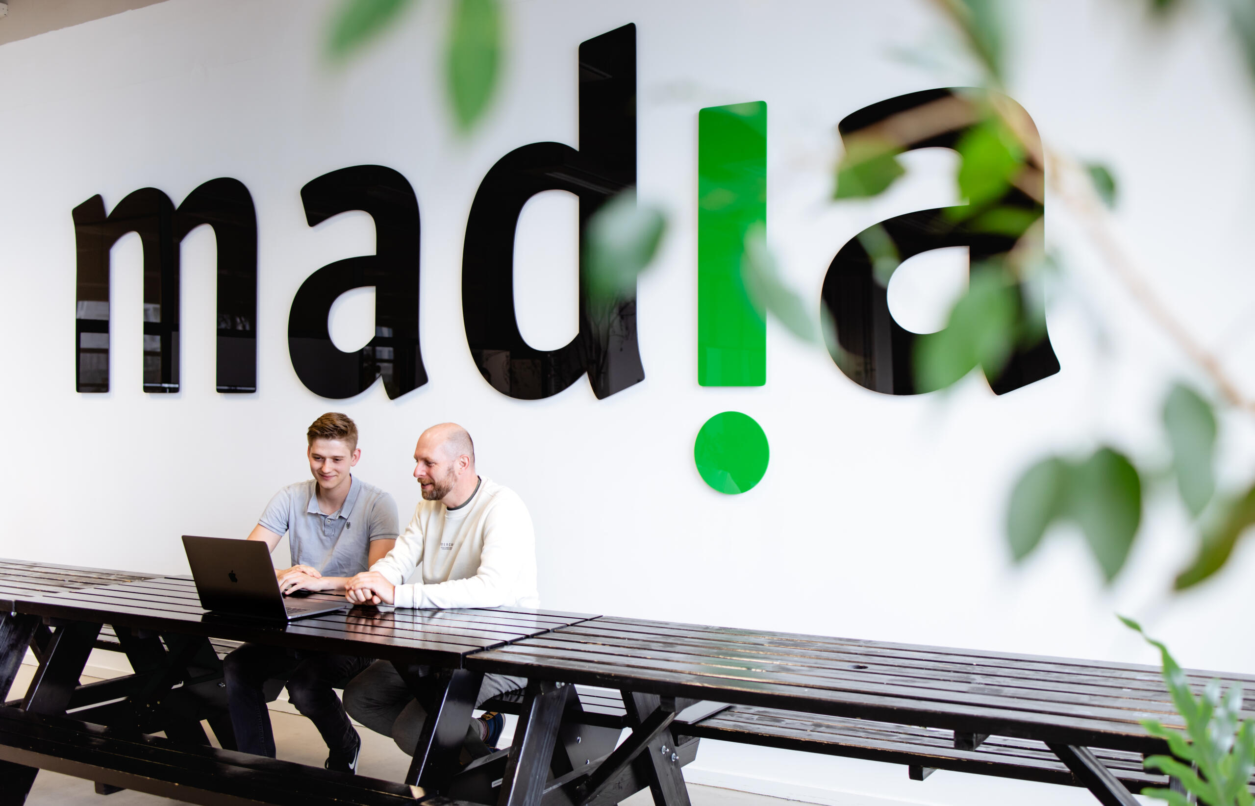 Twee personen werken aan een tafel en ze zitten voor het madia logo