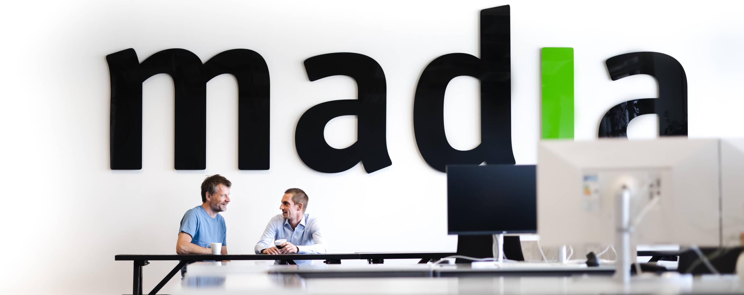 Two people who work at Madia in front of the Madia logo, seated